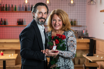 Happy man and woman stand together, man holds in his hand a box with a ring, woman holds bouquet of roses