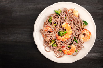A plate of cooked soba, buckwheat noodles, with shrimps and vegetables, shot from above on a dark background with copy space