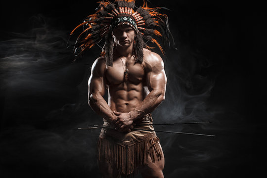 American Indian Apache warrior chief  in traditional clothing and feathered headdress with weapon. Indian chieftain of the tribe with muscled strength body on smoke dark background.