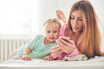 Young mother and her baby daughter using smart phone in bed