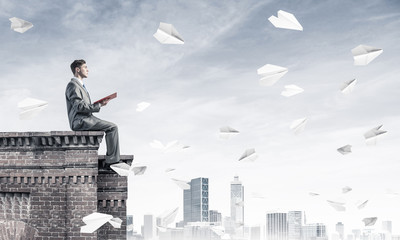 Man on roof edge reading book and cityscape at background