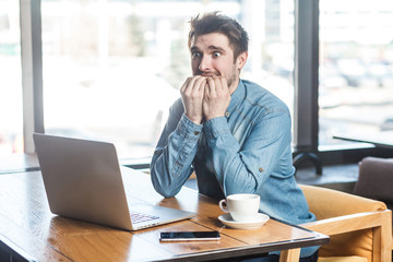 Scared! Side view portrait of emotional nervous young businessman in blue jeans shirt are sitting in cafe, working onlone and nail biting himself cause made big mistake in the laptop. Indoor, inside