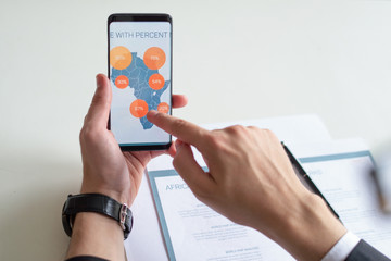Closeup of business man holding smartphone and touching its screen. Manager working with document and diagram. Business analysis concept. Cropped view.