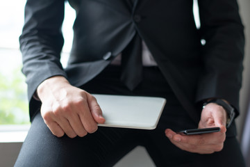 Closeup of business man using digital devices and leaning on sill. Person wearing suit and holding smartphone and tablet computer. Technologies in business concept. Cropped front view.