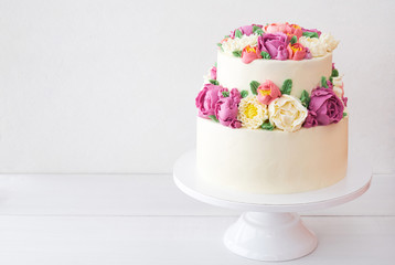 Two-tiered white wedding cake decorated with color cream flowers on a white wooden background.