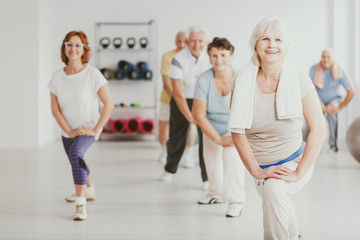 Smiling senior woman exercising with group of active seniors in fitness center - obrazy, fototapety, plakaty