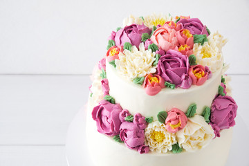 Two-tiered white wedding cake decorated with color cream flowers on a white wooden background.