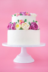 Two-tiered white wedding cake decorated with color cream flowers on a pink background.
