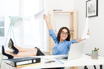 A young brenette girl in a blue shirt and black skirt is sittint at the table in office. She crossed her legs on the table and looks happy and satisfated