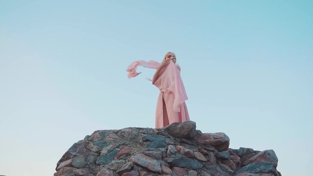 Greek goddess high in a clear blue sky, blonde woman in a chic long pink suit with flying and nagging hem and sleeves posing for the camera, oriental style dresses and fashion