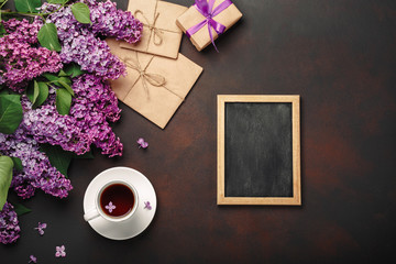 A bouquet of lilacs with cup of tea, chalk board, gift box, craft envelope, a love note on rusty background . Mother's day