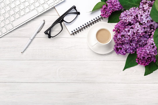 Office desktop with a bouquet of lilacs, coffee cup, keyboard, notebook and pen on white boards