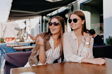 Happy charming caucasian friends expressing positive emotions to camera. Outdoor photo of refined sisters posing on outside cafe. Two joyful cheerful girls have fun while sitting together at cafe