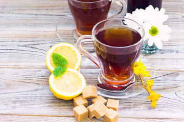 Black tea ceremony - glass full of tea, glass pot, sugar, yellow lemon, flower, tea leaves, spices on a wooden boards background