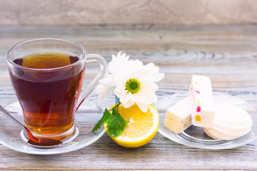 Black tea ceremony - glass full of tea, glass pot, sugar, yellow lemon, flower, tea leaves, spices on a wooden boards background