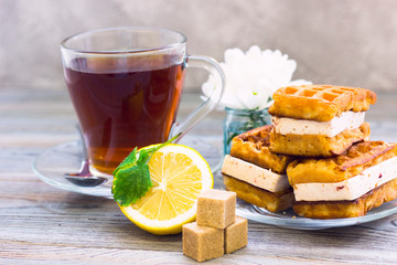 Black tea ceremony - waffles, glass full of tea, glass pot, sugar, yellow lemon, flower, tea leaves, spices on a wooden boards background