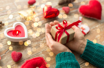christmas, valentines day and holidays concept - close up of female hands holding gift box with heart shaped decorations and candle burning on wooden background