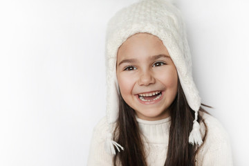 Obraz premium Happy cute child girl in a white winter hat and a white knitted sweater laughs on a white background. Real positive emotions. Smiling beautiful face. 