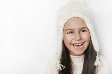 Obraz premium Happy cute child girl in a white winter hat and a white knitted sweater laughs on a white background. Real positive emotions. Smiling beautiful face. 