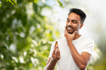grooming and people concept - smiling young indian man applying lotion or beard oil over green...