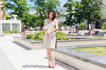 Fototapeta premium Pretty brunette girl in gray dress and vinous heels is standing in park in the city. She holds laptop and looks shy.
