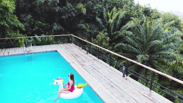 A young beautiful girl with a beautiful figure in a pink swimsuit and sunglasses is resting and tanning in a swimming pool with an inflatable unicorn.