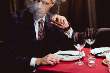 man in suit sitting at table and smoking cigar in restaurant