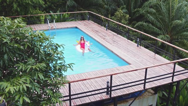 A young beautiful girl with a beautiful figure in a pink swimsuit and sunglasses is resting and tanning in a swimming pool with an inflatable unicorn.