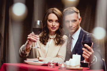 beautiful couple holding glasses of red wine and looking at camera during romantic date in restaurant