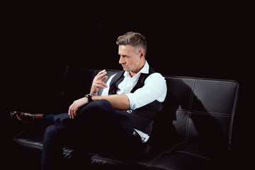 handsome man in formal wear sitting on couch with cigarette isolated on black