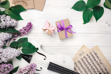 Fototapeta na wymiar A bouquet of lilacs with violin, gift box and music sheet on a white wooden table. Top wiev with space for your text