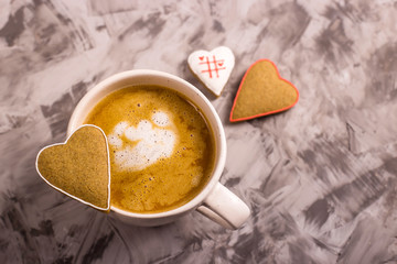 Homemade gingerbread cookies in the shape of a heart with a white outline on a cappuccino cup next to two cookies on a gray background. Valentine's day concept