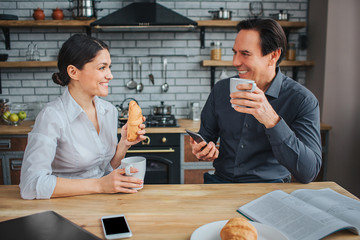 Happy couple sit at table in kitchen. They look at each other. Woman hold croissan and cup in hand. Guy has phone. Happy people smile.