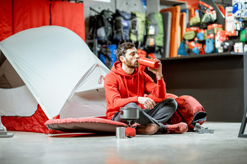 Obraz na płótnie Canvas Man trying camping tools sitting on the floor with travel equipment and tent on the background in the shop