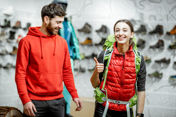 Salesman selling travel equipment to a young woman client trying a backpack in the sports shop