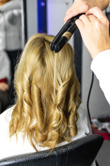 Fototapeta premium Close-up photo professional hairdresser uses flat iron for making curls on beautiful blonde hair. Client sitting on the chair