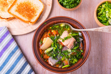 Salmon fillet fish soup with potatoes, tomatoes and carrots sprinkled with green onions and parsley in a glossy plate on a wooden table, top view