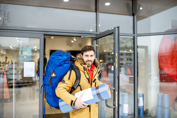 Happy man leaving sports shop with new backpack and camping mat standing in front of the showcase outdoors