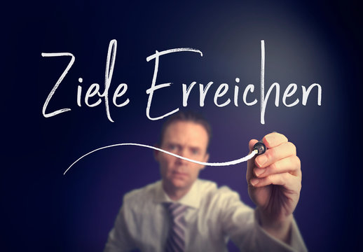 A businessman writing a Achieve Goals "Ziele Erreichen" concept in German with a white pen on a clear screen.