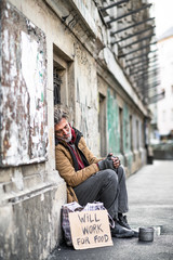 Homeless beggar man sitting outdoors in city asking for money donation.