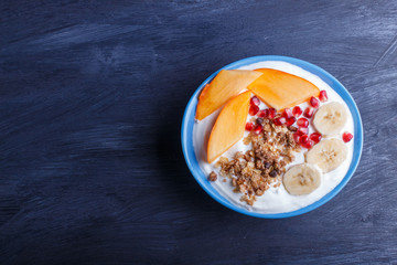 A blue plate with greek yogurt, granola, persimmon, banana, pomegranate on black background.