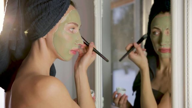 Beauty procedures skin care concept. Young woman applying facial gray and red mud clay mask to her face in bathroom. Woman with different masks on her face multi-masking