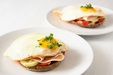 fried egg on wholemeal bread roll with tomato, cucumber and cooked ham, healthy hearty breakfast on a white table, copy space,