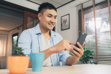 Young Asian man using smartphone while lying on the desk in her living room. Happy male use phone for texting, reading, messaging and buying online at home. Lifestyle man at home concept.