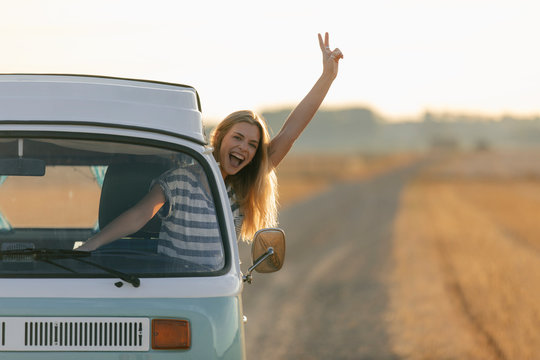 Excited young woman making victory hand sign out of camper van window in rural landscape