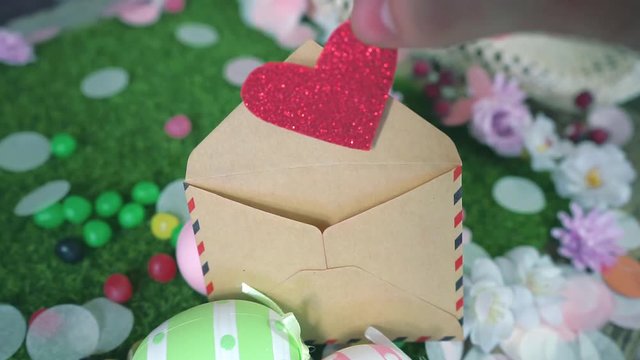 Close-up of hand from a young man hides a glitter red heart into a envelope between Easter eggs in a beautiful spring garden with flowers and candies