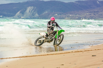 Fototapeta premium Motorcyclist in a protective suit rides a motorcycle on the sea, splashes fly from under the wheels.
