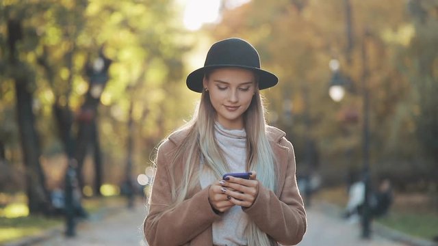 Young beautiful woman wearing stylish coat and black hat using smartphone during walk in autumn city park
