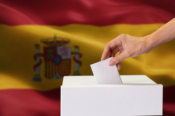 Close-up of human hand casting and inserting a vote and choosing and making a decision what he wants in polling box with Spain flag blended in background.