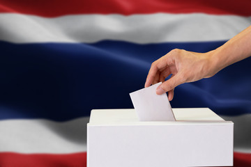 Close-up of human hand casting and inserting a vote and choosing and making a decision what he wants in polling box with Thailand flag blended in background.